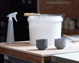 Ein Eimer voller Glasur steht auf einem Tisch. Glasierte Tassen stehen im Vordergrund, weitere Tools sind verteilt.